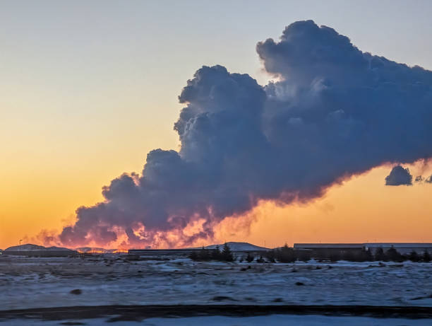 A stunning view of a volcano eruption in Grindavik, Iceland, as seen during sunset. Thick plumes of smoke and ash rise dramatically into the twilight sky, illuminated by the warm glow of the setting sun. Snow-covered ground adds contrast to the fiery display.