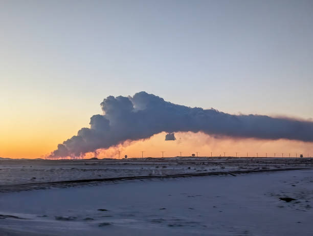 A stunning view of a volcano eruption in Grindavik, Iceland, as seen during sunset. Thick plumes of smoke and ash rise dramatically into the twilight sky, illuminated by the warm glow of the setting sun. Snow-covered ground adds contrast to the fiery display.