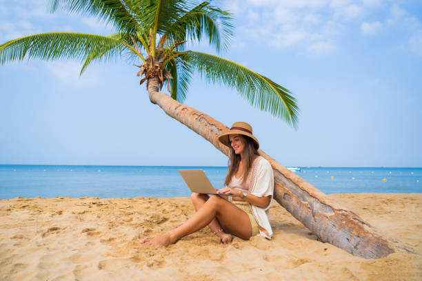 istock Young satisfied happy smiling nomad woman freelancer sitting under a palm tree and working remotely online at a laptop, enjoying a dream job anywhere in the world 2161755390