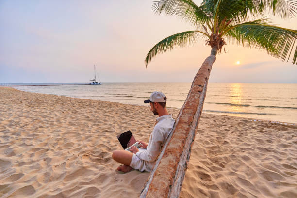 istock Young nomad man freelancer sitting under a palm tree and working remotely online at a laptop, enjoying a dream job anywhere in the world 2161763448