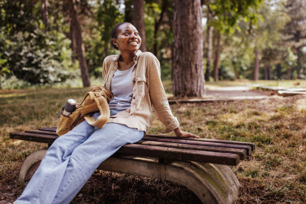 A person is leisurely leaning back on a wooden park bench surrounded by a natural green environment, soaking up the sun and enjoying a moment of relaxation amidst the trees.
