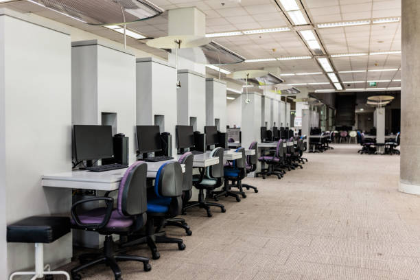 Interior view of a modern Bangkok university library. Rows of empty workstations, each equipped with an office chair and a desktop computer, stretch across the area.