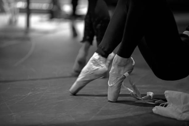 Young female ballerina legs in white tulle skirt stands elegantly on wooden floor. Soft lighting highlights poised feet in satin pointe shoes, creating serene atmosphere