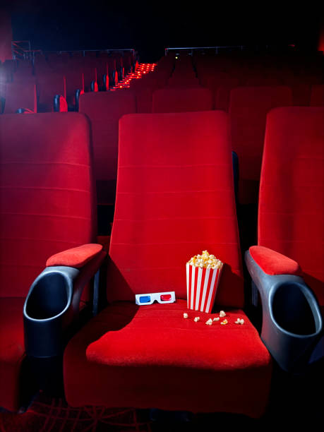 Image of striped carton of popcorn and 3D glasses in cinema theatre seat surrounded by spilled popped kernels, disposable drink cup and drinking straw in cup holder, rows of empty, red velvet cinema seats, focus on foreground stock photo