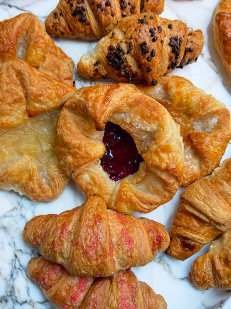 Close-up image of assorted, freshly baked breakfast pastries on marble counter, raspberry and apricot jam filled pastries, chocolate hazelnut, vegan raspberry and all butter croissants, elevated view
