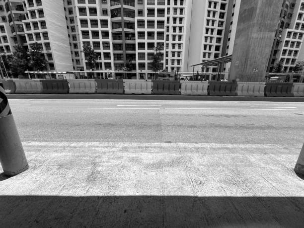 A black and white photograph of an urban street scene featuring construction barriers, high-rise buildings, and concrete surfaces, highlighting the modern city environment.