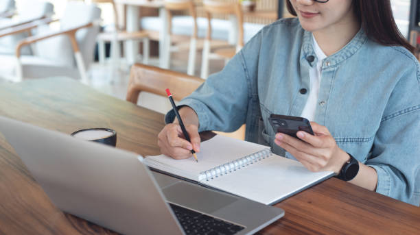 istock Young asian business woman taking note on notebook, using mobile phone and working on laptop computer at coffee shop. Student studying online, writing on notebook, doing homework, e-learning, remote work 2161834362