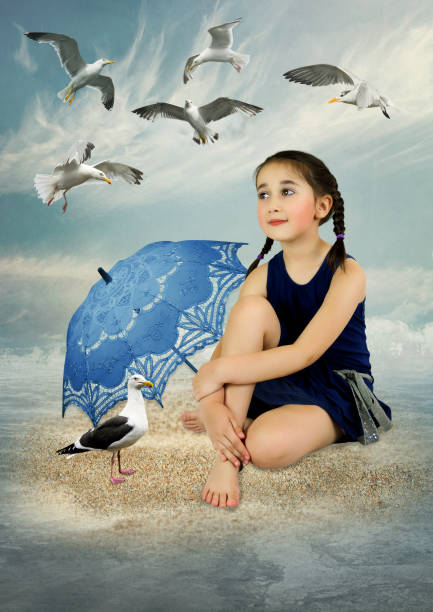 A little girl sitting on the sea sand under a blue lacy umbrella looks with tenderness at the fluttering seagulls