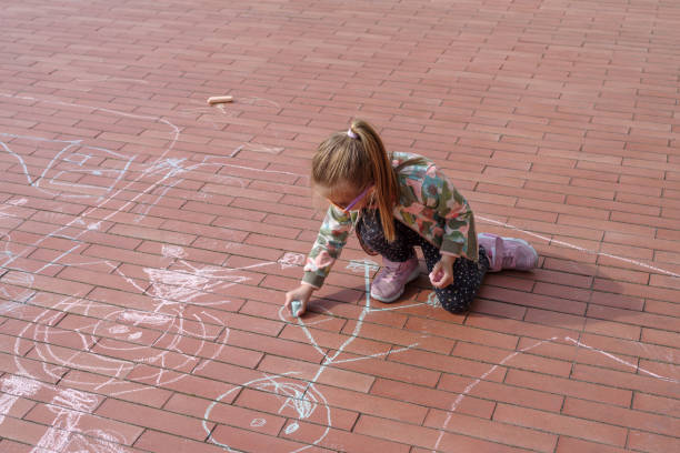 Little preschool girl painting rainbow with colorful chalks on ground on backyard. Positive happy toddler child drawing and creating pictures. Creative outdoors activity in summer