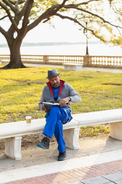 A senior African-American man in his 60s sitting on a bench in a city park reading a book. He is wearing a jacket and hat, with a cup of coffee to stay warm on a chilly autumn day.