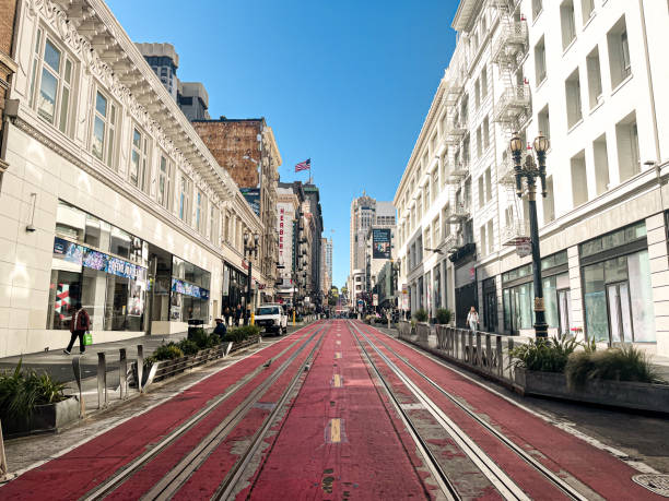 View up to the top of the hill in San Francisco. Middle of the road painted in red, tram railings going far up the road. Cozy city architecture around.