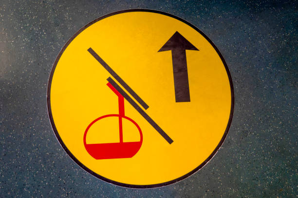 A bright yellow floor sign in the departure building of a mountain cable car. It is directing passengers towards the boarding area, with an arrow that points straight ahead. The red image of a cable car attached to cables, gives people information on which way to go.