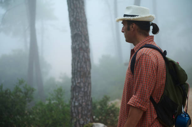 Hiker in a misty forest wearing a straw hat and checkered shirt, carrying a backpack. Foggy atmosphere creates a mysterious and serene environment.