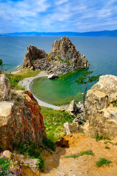Cove on Lake Baikal with a curved beach, rocky cliffs, and transparent emerald-blue water under a partly cloudy sky