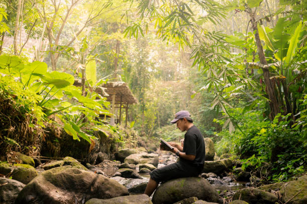 istock Young Indonesian Man Enjoying Reading Sitting on Large River Rock 2161938868