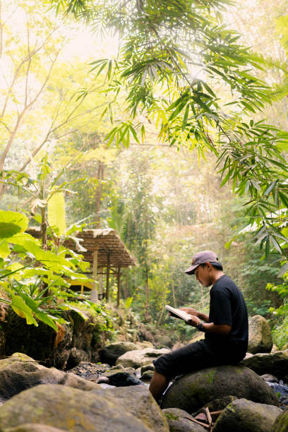 istock Young Indonesian Man Enjoying Reading Sitting on Large River Rock 2161938905