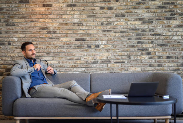 istock Pensive male developer holding eyeglasses and looking away seriously while relaxing on sofa at his workplace 2161981220