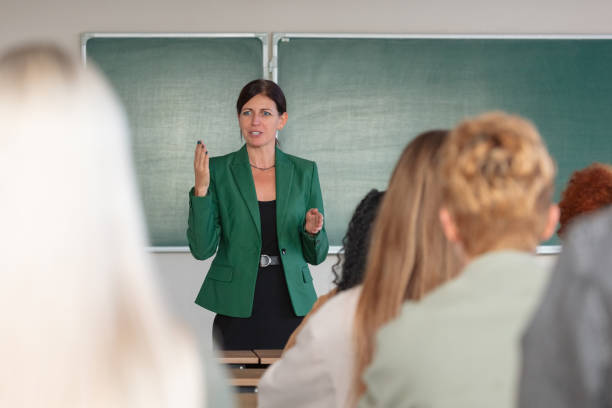 Female teacher wearing green jacket standing in front of blackboard in the classroom and talking to university students.