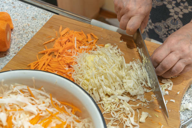 The cook thinly cuts white cabbage with a knife on a wooden cutting board to prepare a vegetable salad with carrots. Veganism and raw food diet concept.