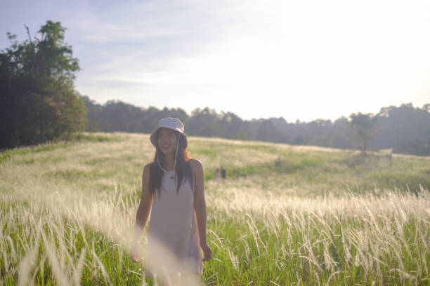 Young woman in a straw hat and floral dress pointing upwards in a sunlit park setting.