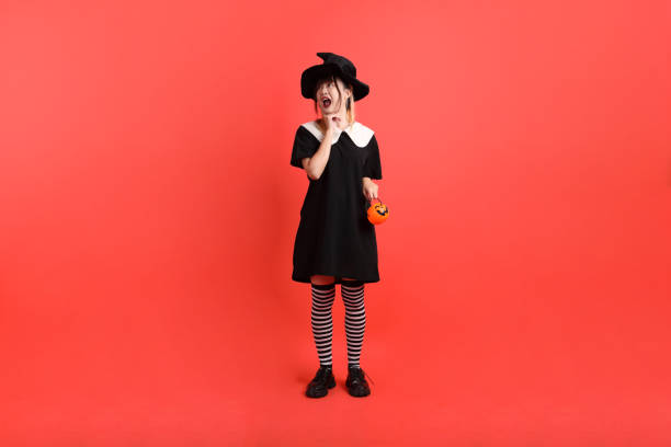 Portrait of one Caucasian beautiful teenage girl in a witch hat with tulle, who creatively plays with Halloween eye candies, putting them to her eyes with her mouth open, on an autumn sunny morning, close-up side view. Happy Halloween concept, family celebration, holiday breakfast time.