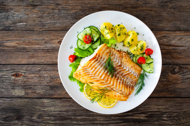 istock Fried cod fillet, boiled potatoes, fresh vegetables and lemon slices served on white plate on wooden table 2162011022