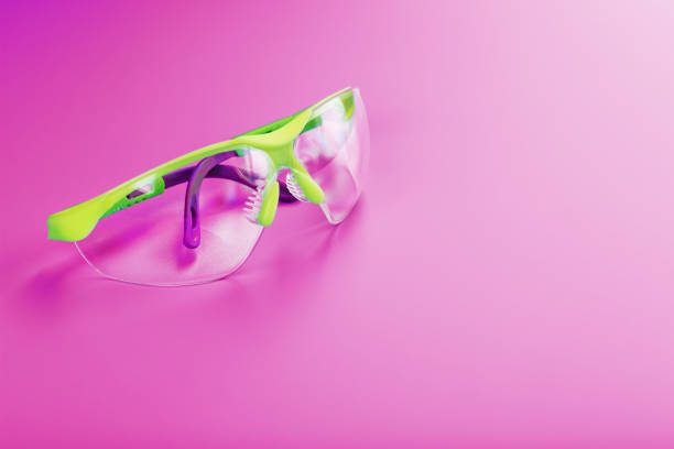 Protective safety glasses rest on an outdoor table surface, illuminated by a warm sunlit background with soft-focus natural scenery. Represents safety, outdoor work, and the sunny environment.
