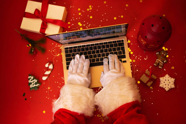 istock Santa Claus with laptop. Hands in white gloves typing on an notebook keyboard. Gift boxes, christmas cookies, lantern and fir branch on red background. 2162027779