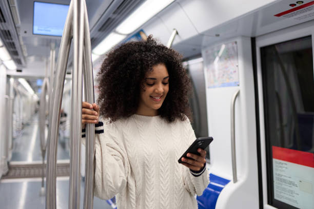 istock Young multiethnic woman is standing on a subway train, holding onto a railing and smiling while using her smartphone 2162030227