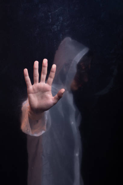 istock Young man wearing a transparent plastic raincoat behind glass making hand gestures at the camera. Face hidden. Studio portrait. 2162037271