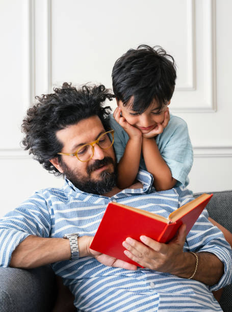 istock Father and son sitting on sofa at home and reading book together 2162046717
