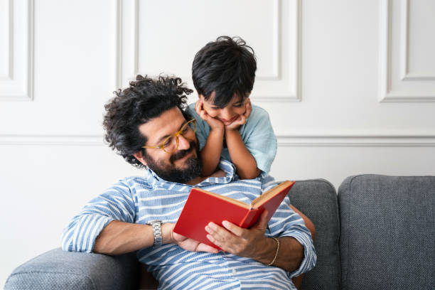 Father and son sitting on sofa at home and reading book together