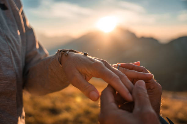 In the scenic outdoors of the mountains, a mature man shares a moment of profound connection, holding his partner's hand and slipping on an engagement ring during a romantic proposal.
