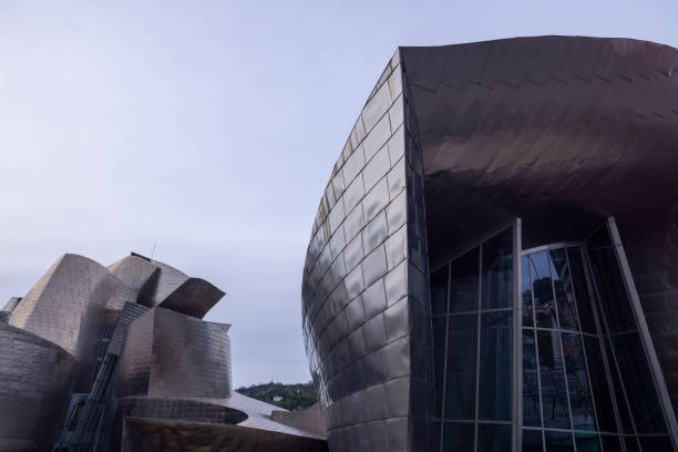 The Guggenheim Museum in Bilbao stands proudly by the riverside, its titanium curves glistening in the daylight. The iconic structure, designed by Frank Gehry, reflects the sunlight, creating a dazzling display of modern architecture. The museum's unique forms and the tranquil water of the Nervión River blend harmoniously, offering a striking visual contrast against the blue sky. Visitors stroll along the promenade, admiring the museum's innovative design and its integration with the natural surroundings