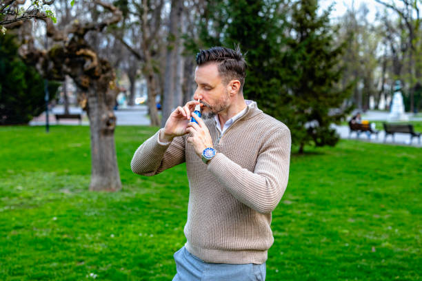 A young man wearing a tan sweater and blue pants stands in a park on a sunny day. He is using a nasal spray to help with his allergies. He has a slight beard and short brown hair. The grass and trees in the background are green and lush.