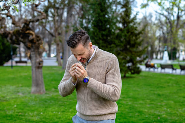A young man stands in a park with trees and green grass in the background. He is wearing a beige sweater and is holding a tissue to his nose, sneezing. He appears to be experiencing allergies.