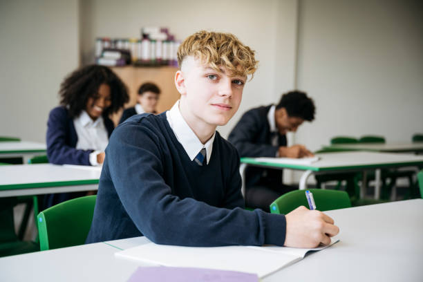 Portrait of British secondary schoolboy taking exam Waist up view of blond teenager in uniform sitting at desk and pausing from writing in booklet to look at camera with slight smile, classmates in background. teenager boy haircuts stock pictures, royalty-free photos & images