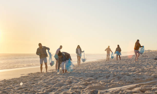 People, group and cleaning for garbage at beach for recycling, trash and plastic for marine ecology. Community service, men or women with volunteering, sustainability or environment by sea at sunset