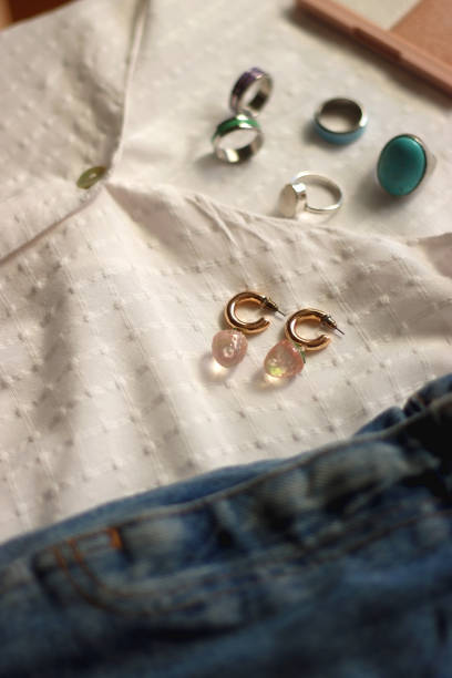 White blouse, denim skirt, colorful rings, earrings and beauty products on white background. Summer outfit in pastel colors. Selective focus.