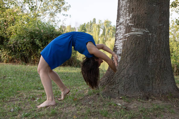 Young woman wearing a blue dress is doing a backbend yoga pose by a tree in a park