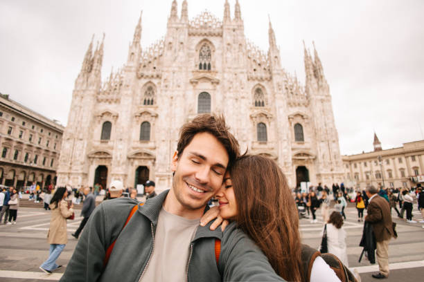 Young couple stands closely together, smiling and posing for a photo in front of an architectural background. The man is wearing a light beige shirt, while the woman sports a white shirt with a necklace and a shoulder bag. Their expressions are joyful, with the woman showing her curly hair and the man standing confidently with his arm around her. The image captures a moment of happiness and togetherness in an outdoor setting.