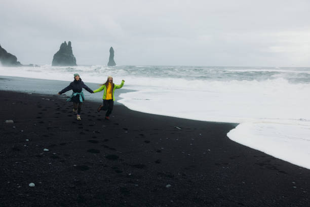Front view of two cheerful female explorers holding hands and feeling freedom running at majestic Reynisfjara black sand beach in Vik, South Iceland contemplating the views of natural arch, cliffs and beautiful stormy ocean