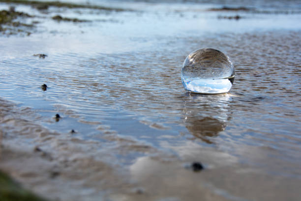 A glass ball lies in the waves on the sandy beach, the sea and the setting sun are reflected in the ball