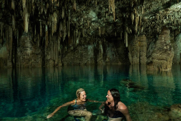 Two women in swimsuits swim in a mysterious cenote with greenish water and limestone walls in an underground cave, Yucatan, Mexico