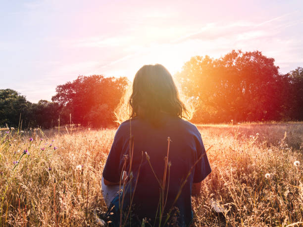 Individual sitting in a meadow, facing the sunset, surrounded by tall grass and trees, enjoying nature's tranquility.