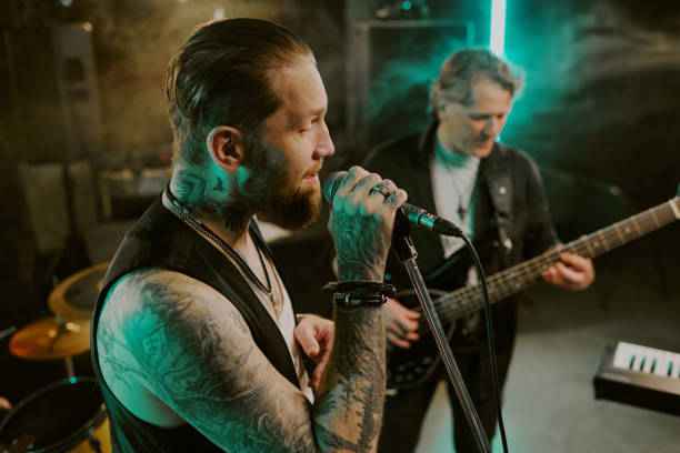 Medium closeup of bearded rock vocalist singing song on stage, mature man playing bass guitar in background