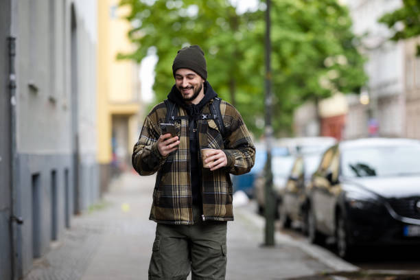 istock Smiling young man wearing hat and jacket walking on the street using cell phone 2162141935