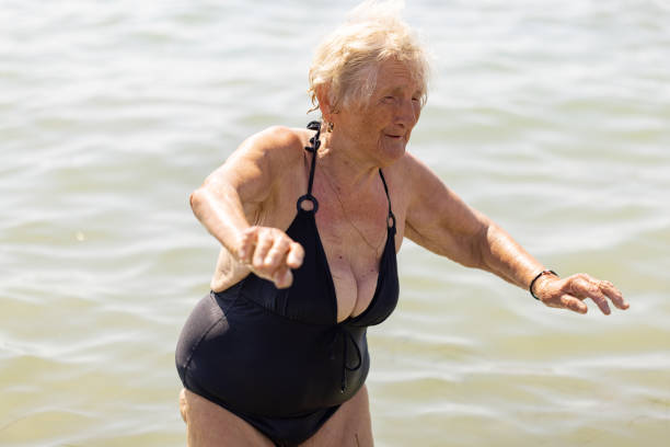 A senior lady in a black bathing suit seeks support as she steps out of the ocean and onto the shore.