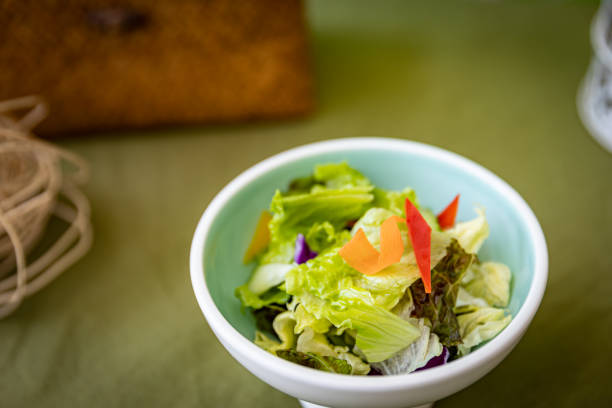 An assortment of bright green lettuce leaves are neatly arranged in a pale white bowl, accented by diced orange carrots and red pepper slices that add dynamic visual interest. The fresh, crisp texture of the vegetables promises a light and healthy eating experience. This meticulously prepared salad not only offers delicious and nutritious flavors, but also presents an elegant and visually appealing display.