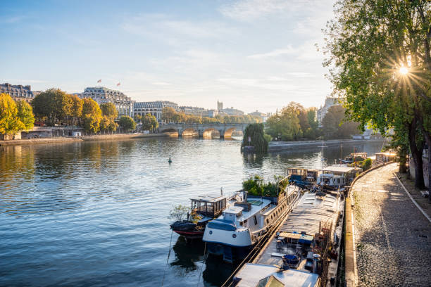 Early morning light shines over the Seine River in Paris, France, illuminating boats docked along the tree-lined promenade. The historic architecture and bridges add charm to this serene urban landscape.